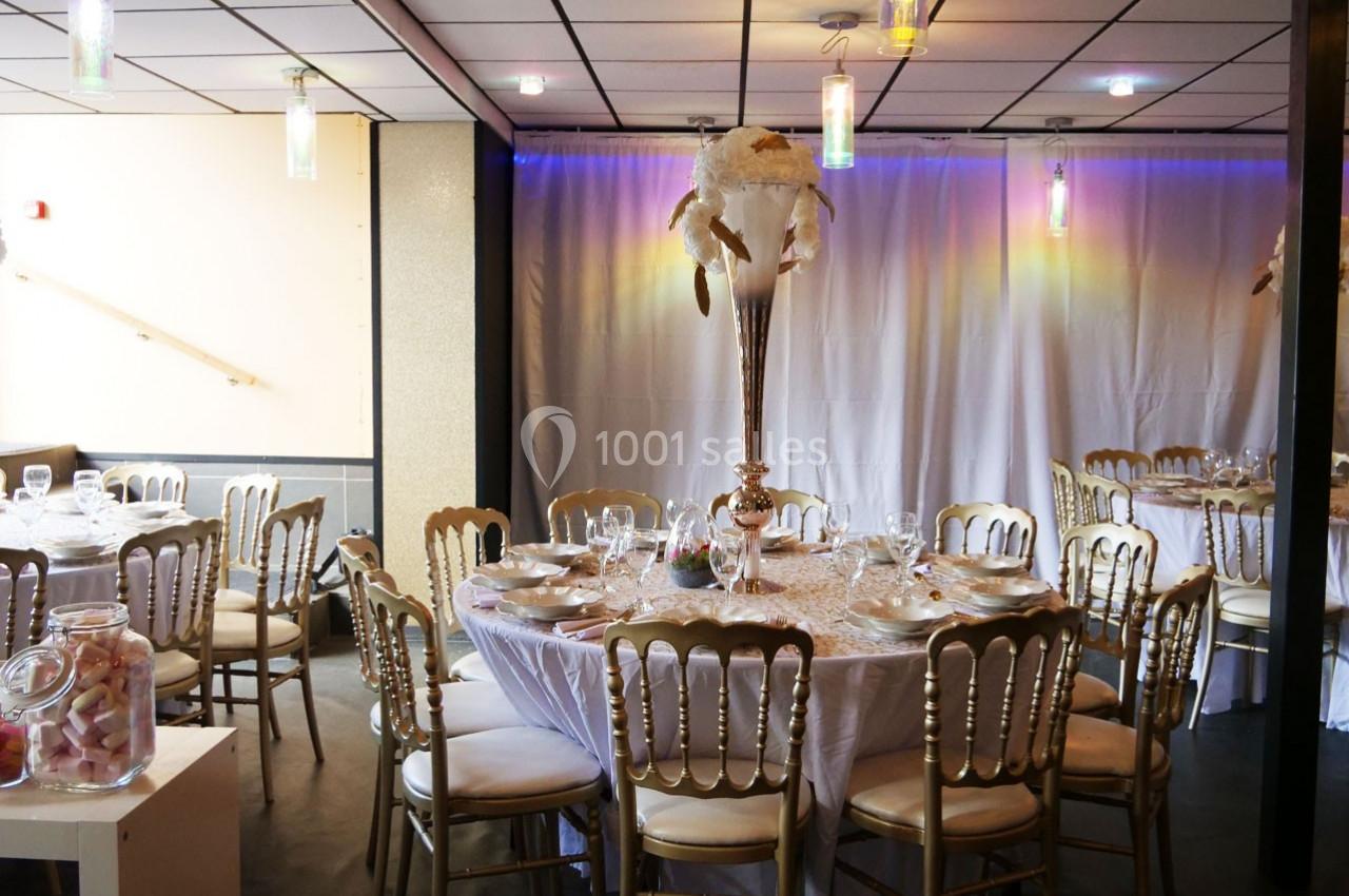 Salle de réception décorée avec des tables rondes, nappes blanches, chaises dorées et un centre de table floral.