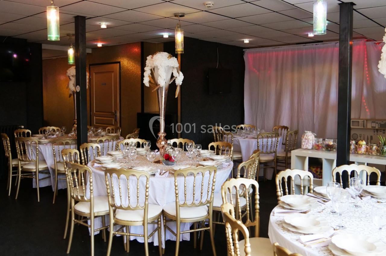 Salle de réception décorée avec des tables rondes dressées, chaises dorées et centre de table floral blanc.