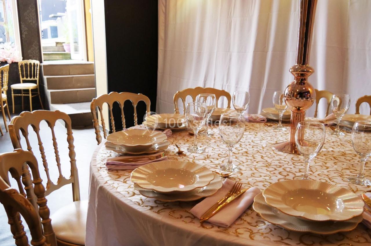 Table ronde élégamment dressée avec nappe blanche et dorée, assiettes en porcelaine, verres à pied et chaises dorées.