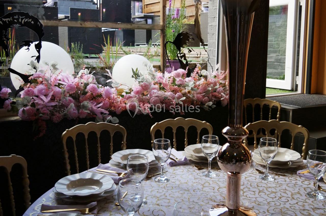 Table dressée avec vaisselle élégante, centre de table argenté et décor floral rose près d'une baie vitrée.