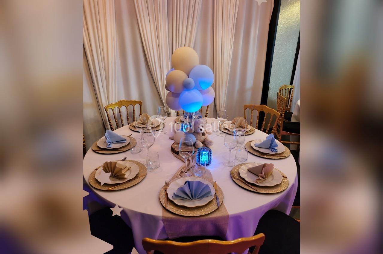 Table décorée pour un événement avec nappes blanches, serviettes pliées, ballons et un ours en peluche au centre.