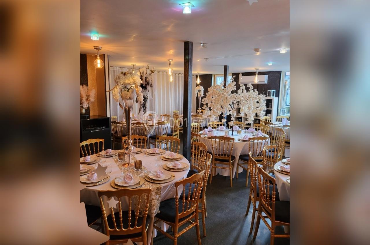 Salle décorée pour un événement avec tables rondes, chaises dorées et décorations florales blanches élégantes.