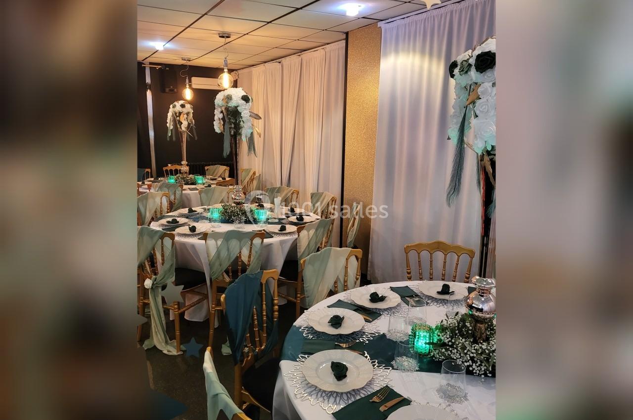 Salle décorée pour un événement avec tables rondes dressées, nappes blanches, chaises dorées et centres de table fleuris.