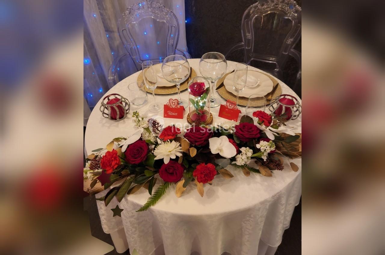 Table ronde élégamment dressée avec nappes blanches, décor floral rouge et blanc, verres et assiettes dorées.