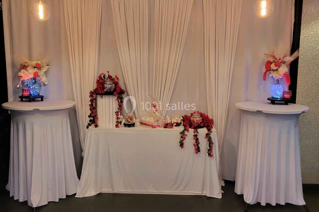 Décoration de table avec nappes blanches, fleurs rouges et roses, et éléments décoratifs sous un fond de rideaux blancs.