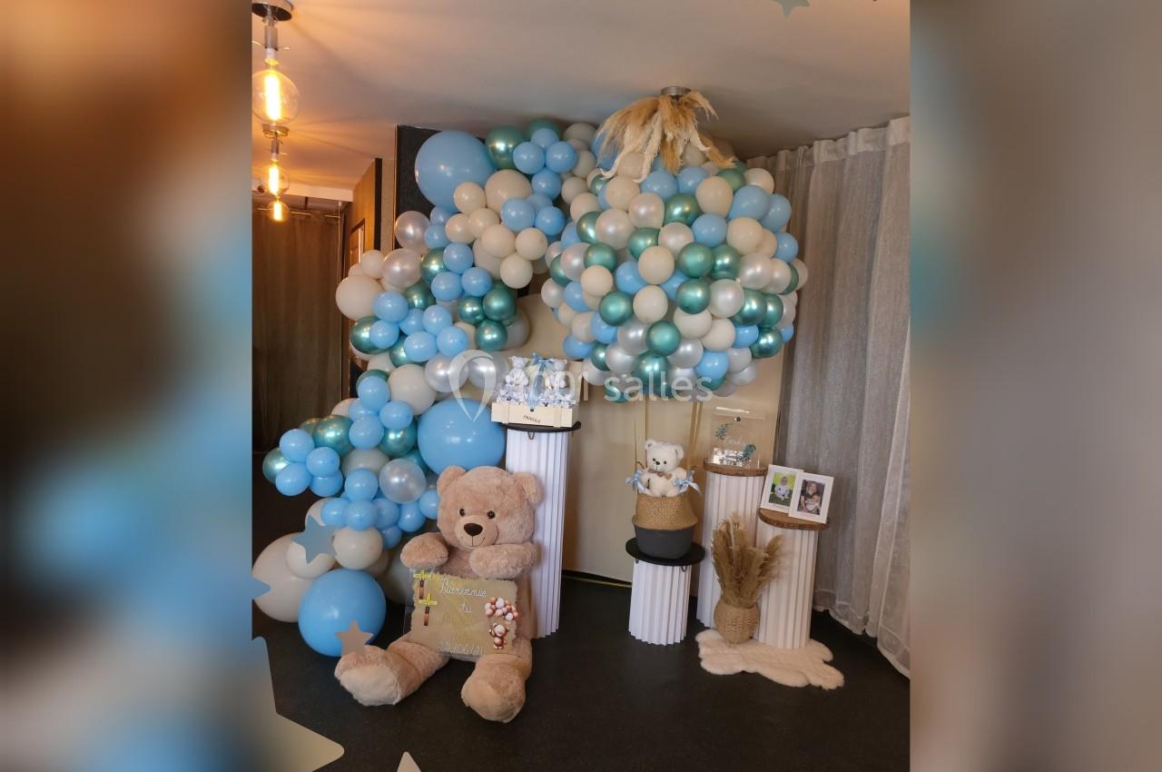 Décoration festive avec arche de ballons bleus et beiges, peluches, et éléments décoratifs sur des colonnes blanches.