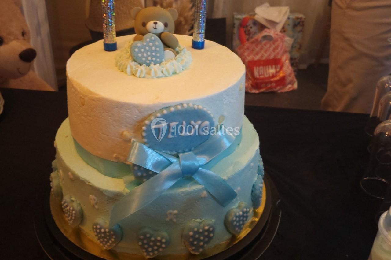 Gâteau à deux étages décoré de bleu et blanc, avec un ours en figurine, des cœurs et le prénom ’Telyo’.