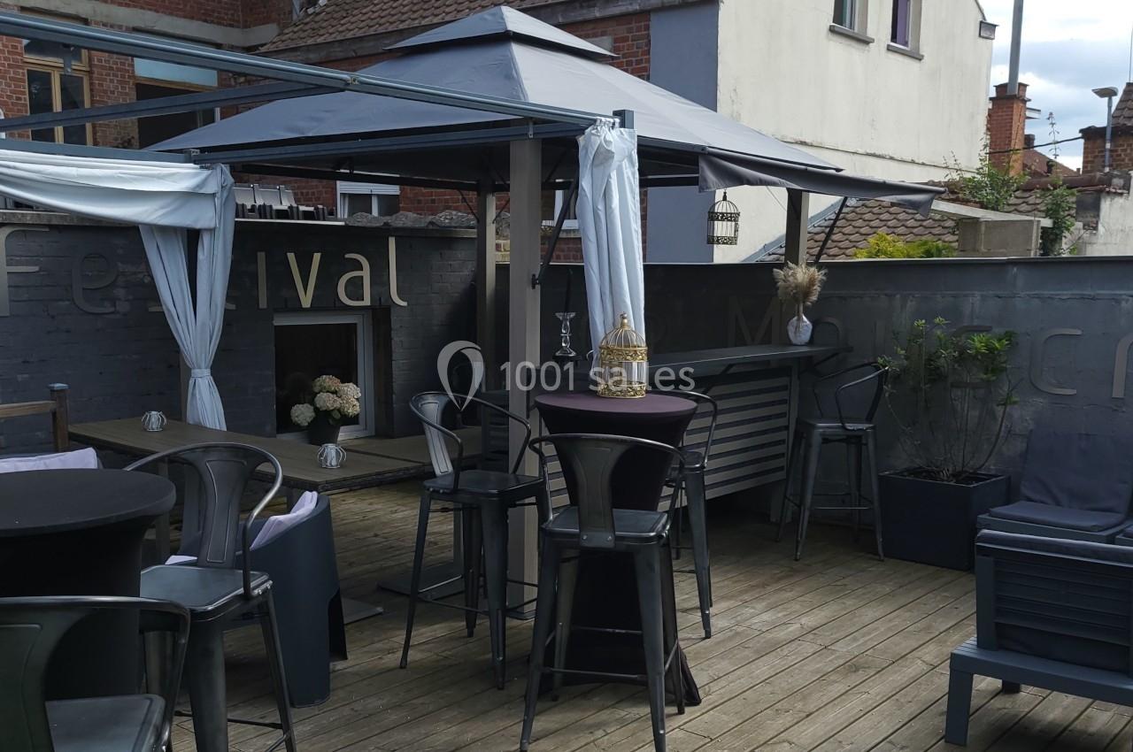 Terrasse en bois avec tables hautes, chaises en métal, pergola et décorations, entourée de murs gris.