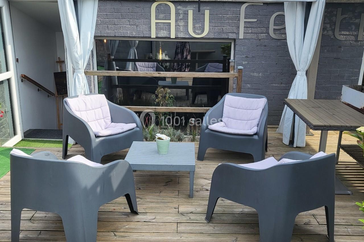 Quatre fauteuils gris autour d'une table basse sur une terrasse en bois, avec une façade en arrière-plan.