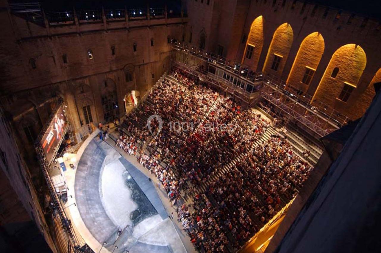 Vue d'une scène de théâtre en plein air, entourée de gradins remplis de spectateurs, dans un cadre architectural historique.