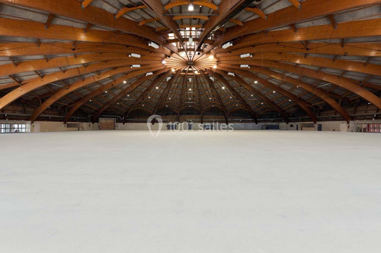Vue intérieure d'une grande salle avec une structure en bois voûtée et un sol clair, éclairée par des fenêtres en hauteur.