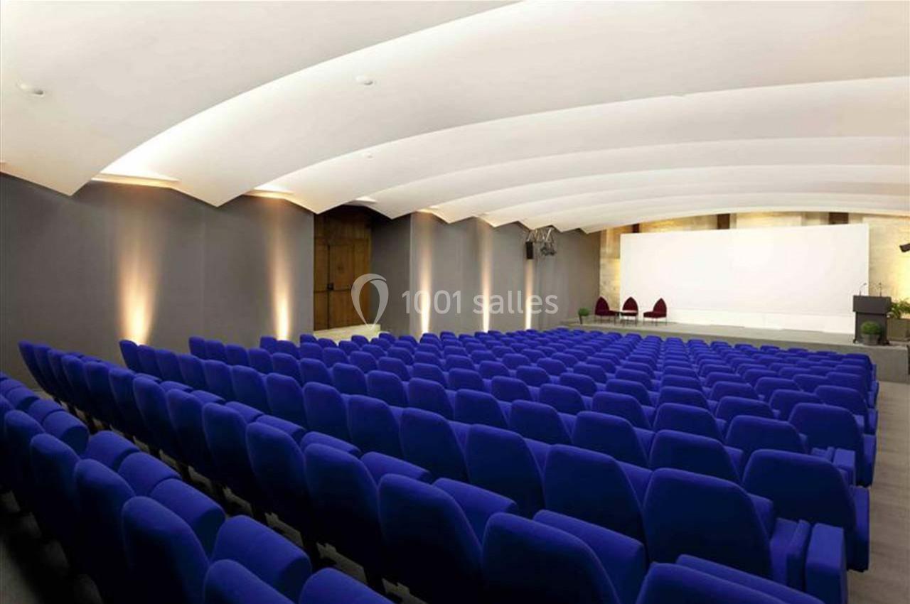 Salle de conférence avec rangées de sièges bleus, éclairage tamisé et écran blanc en fond.