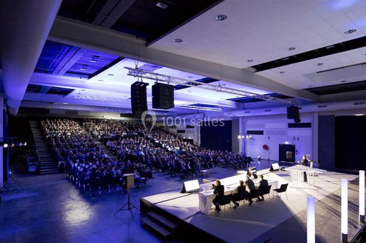 Salle de conférence avec un public nombreux, une scène éclairée et un orateur s'exprimant au micro.