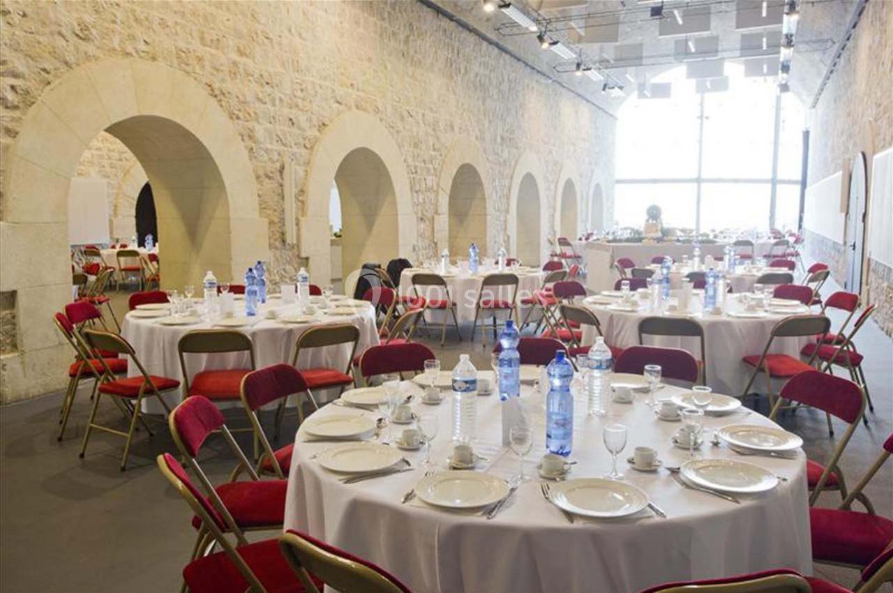 Salle en pierre avec des tables rondes dressées, chaises rouges, vaisselle blanche et bouteilles d'eau, éclairée par une…