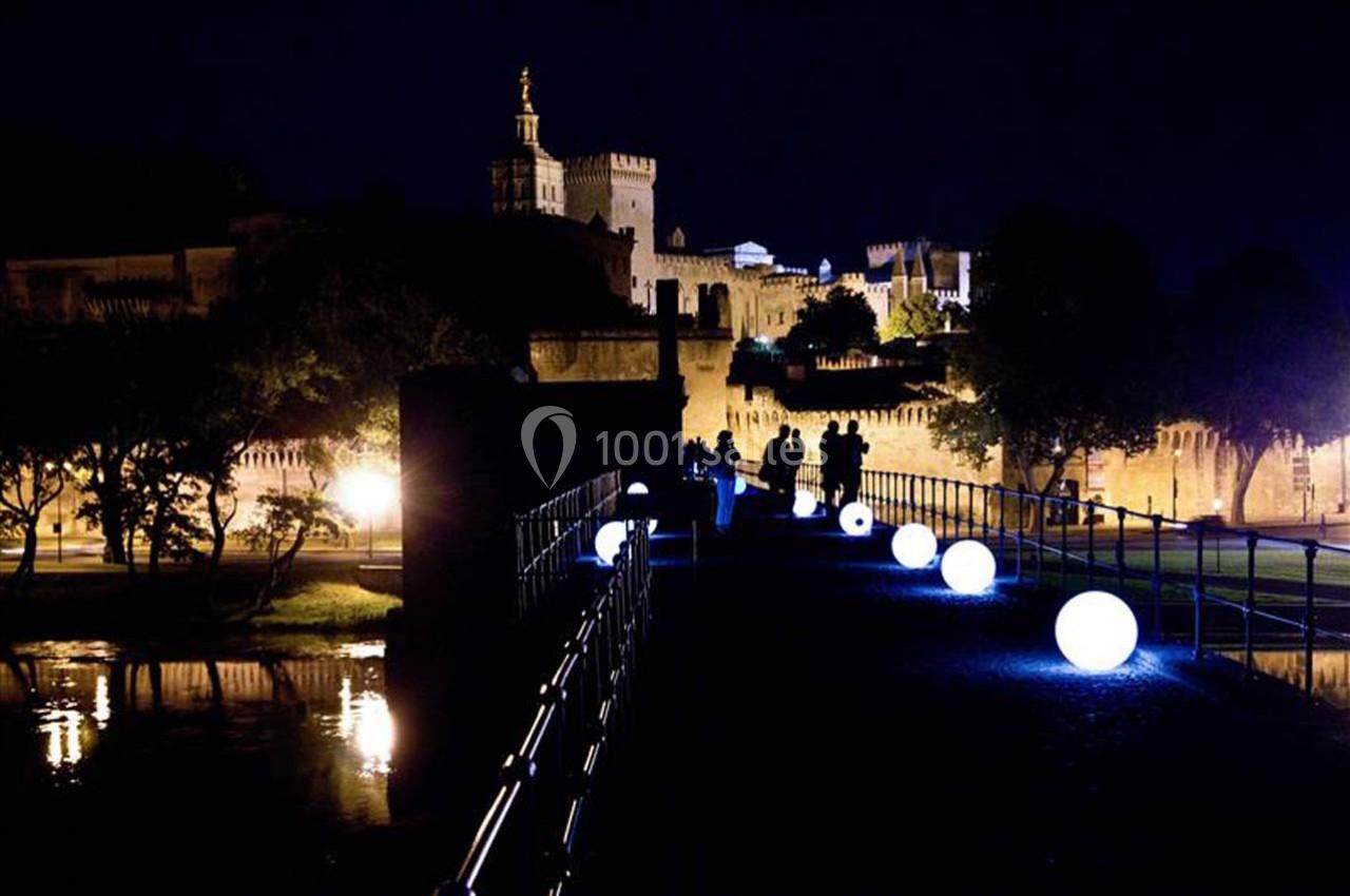 Passerelle illuminée par des sphères lumineuses menant à un bâtiment historique éclairé de nuit.