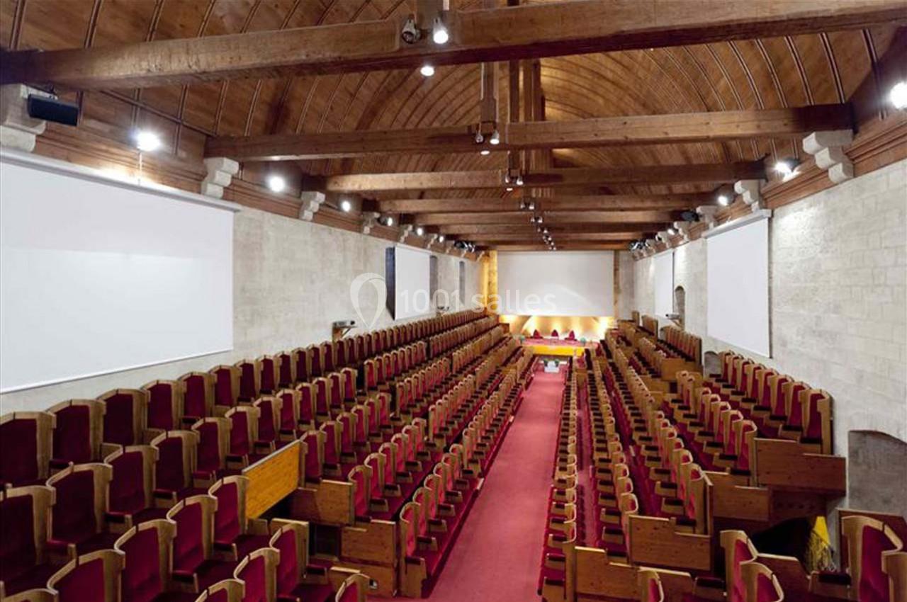 Salle de conférence avec rangées de sièges en bois et velours rouge, plafond voûté en bois et murs en pierre.