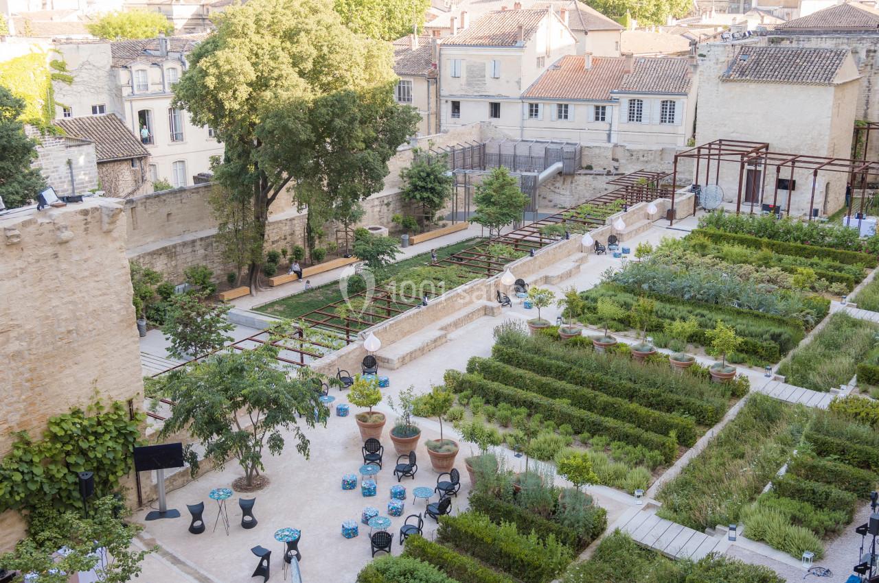 Vue aérienne d'un jardin aménagé avec des plantes, des allées, des tables et des bâtiments en arrière-plan.