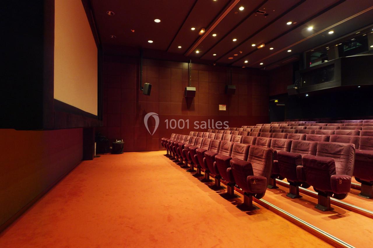 Salle de cinéma vide avec des rangées de fauteuils rouges et un écran blanc sur la gauche.