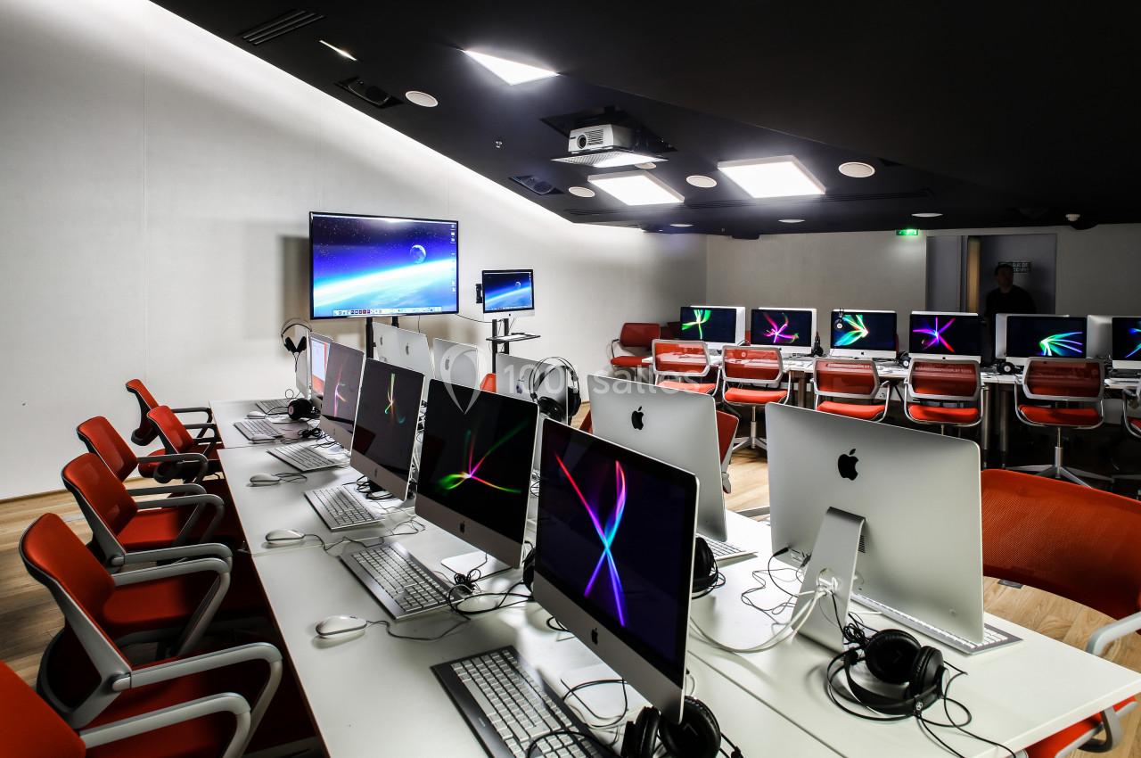 Salle informatique moderne avec des ordinateurs Apple alignés sur des bureaux, chaises rouges et écran mural allumé.