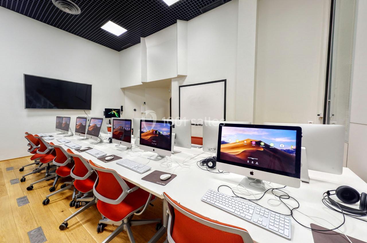 Salle équipée d'ordinateurs iMac alignés sur des bureaux blancs avec des chaises rouges, écran mural et tableau blanc.