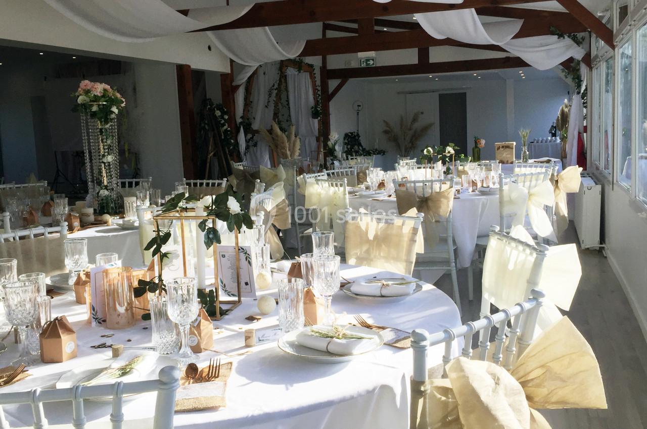 Salle de réception décorée pour un événement, avec tables rondes dressées, nappes blanches et ornements floraux.