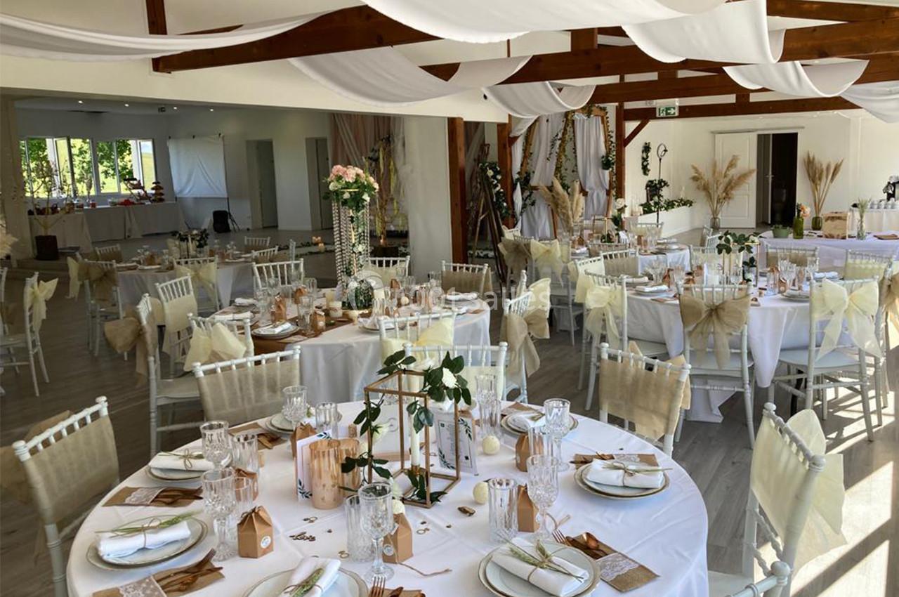 Salle de réception décorée pour un événement, avec tables rondes dressées, nappes blanches et ornements floraux.