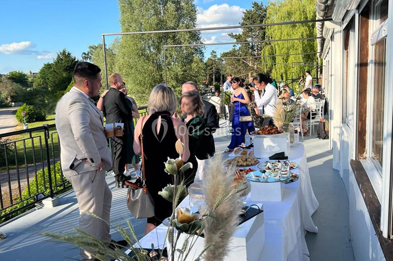 Groupe de personnes discutant sur une terrasse ensoleillée, avec des tables dressées pour un buffet en extérieur.