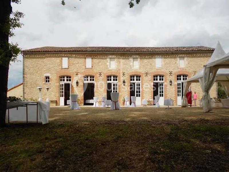 Façade d'un bâtiment en pierre avec grandes fenêtres, tables dressées à l'extérieur et tente blanche sur la droite.