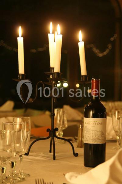 Chandelier allumé et bouteille de vin rouge sur une table dressée avec verres et nappes blanches.