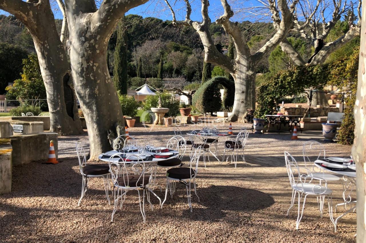Terrasse extérieure avec tables et chaises en fer forgé sous de grands arbres, entourée de végétation et de pots décoratifs.