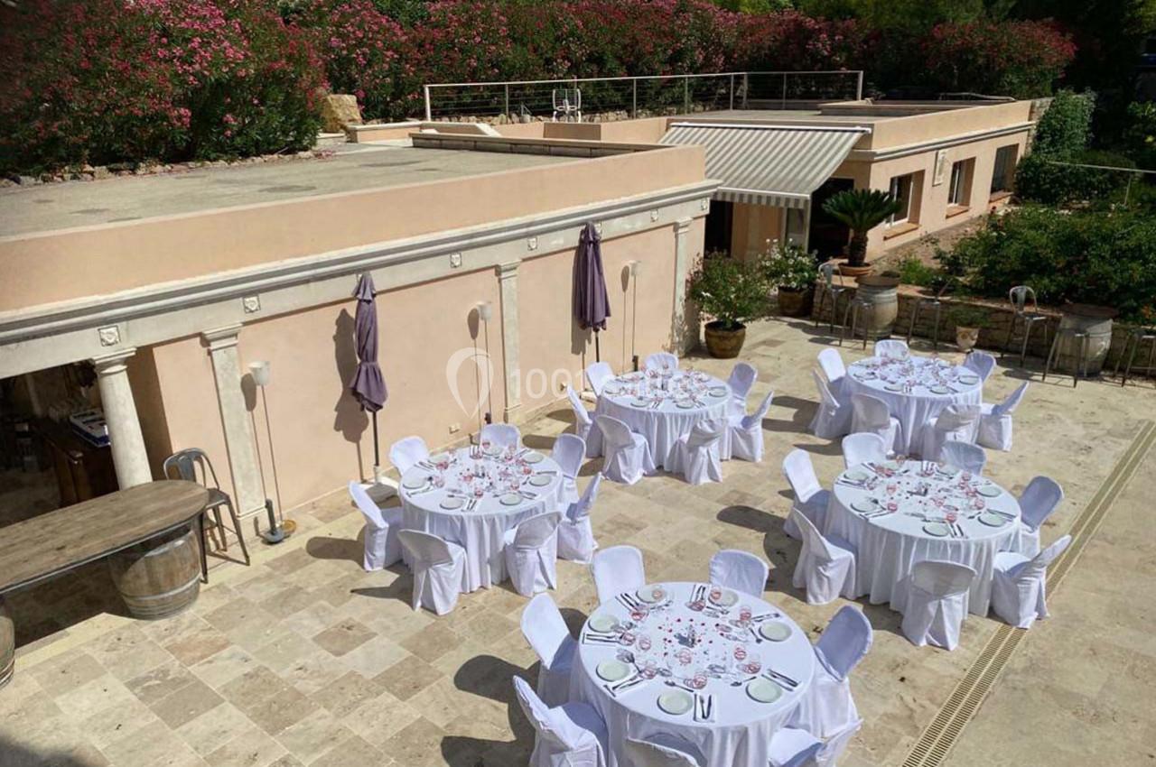 Tables rondes dressées avec nappes blanches sur une terrasse en pierre, entourée de verdure et de bâtiments clairs.