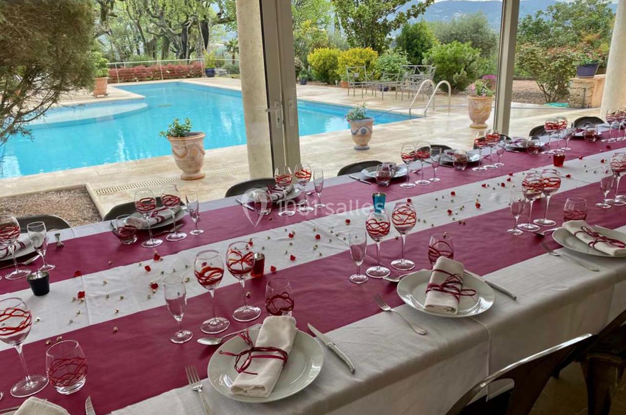Table dressée avec nappes rouges et blanches, couverts et verres, près d'une piscine entourée de végétation.