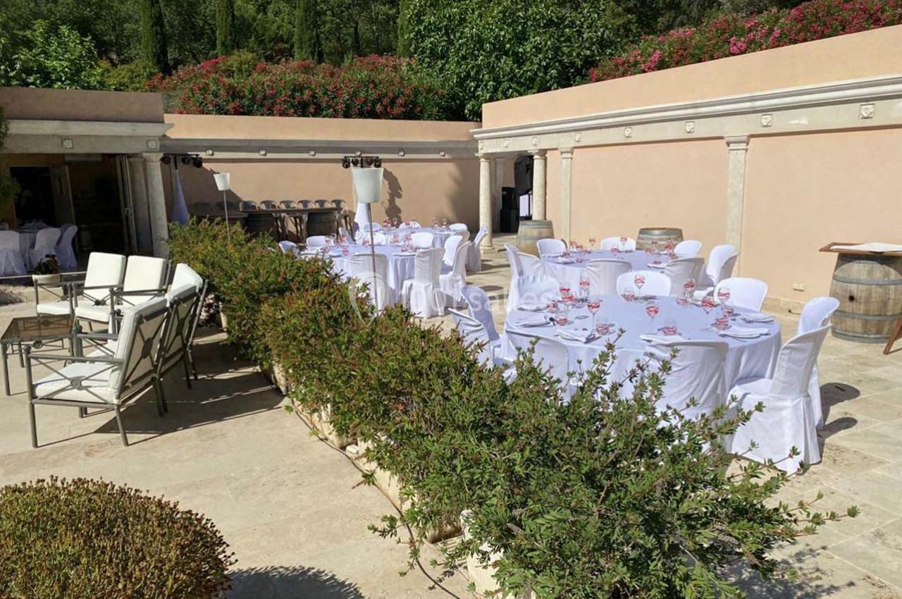 Terrasse extérieure avec tables rondes dressées et chaises blanches, entourée de verdure et de murs en pierre beige.