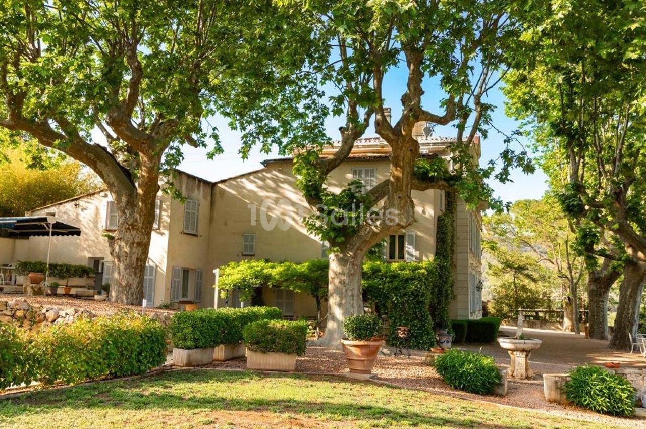 Grande maison en pierre entourée d'arbres, avec terrasse ombragée et pots de fleurs dans une cour ensoleillée.
