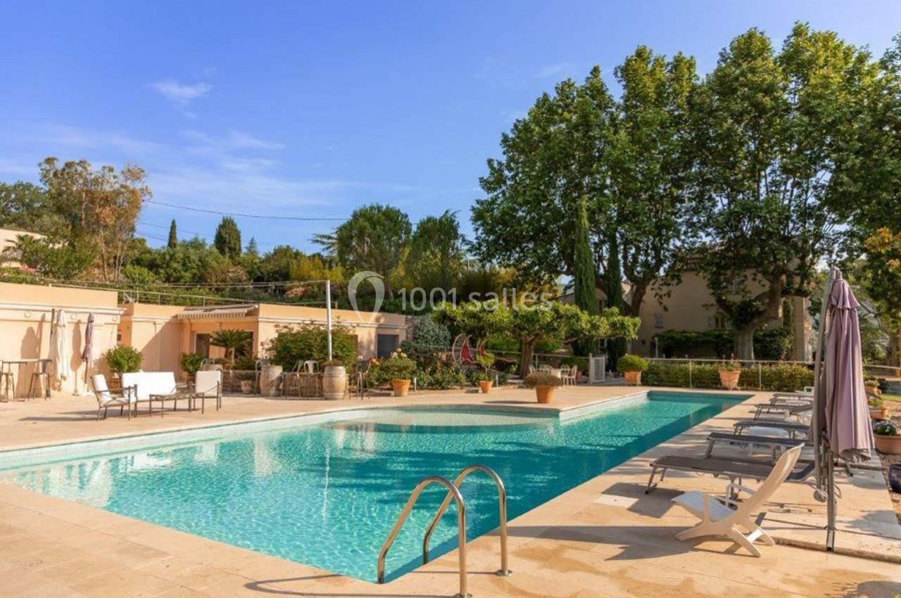 Piscine extérieure entourée de chaises longues, parasols et végétation, sous un ciel bleu clair.