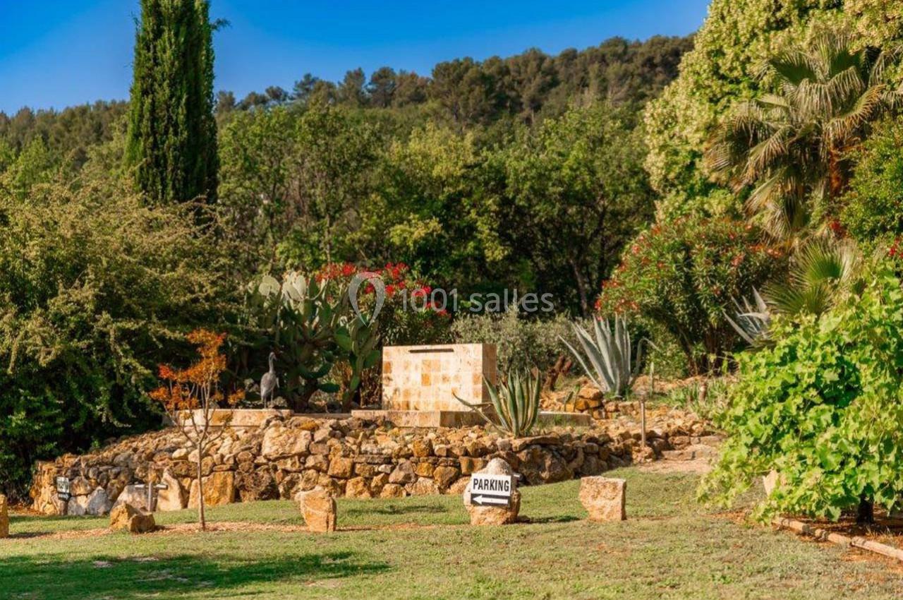 Jardin méditerranéen avec cactus, arbres et arbustes, muret en pierre, panneau ’Parking’ et collines en arrière-plan.