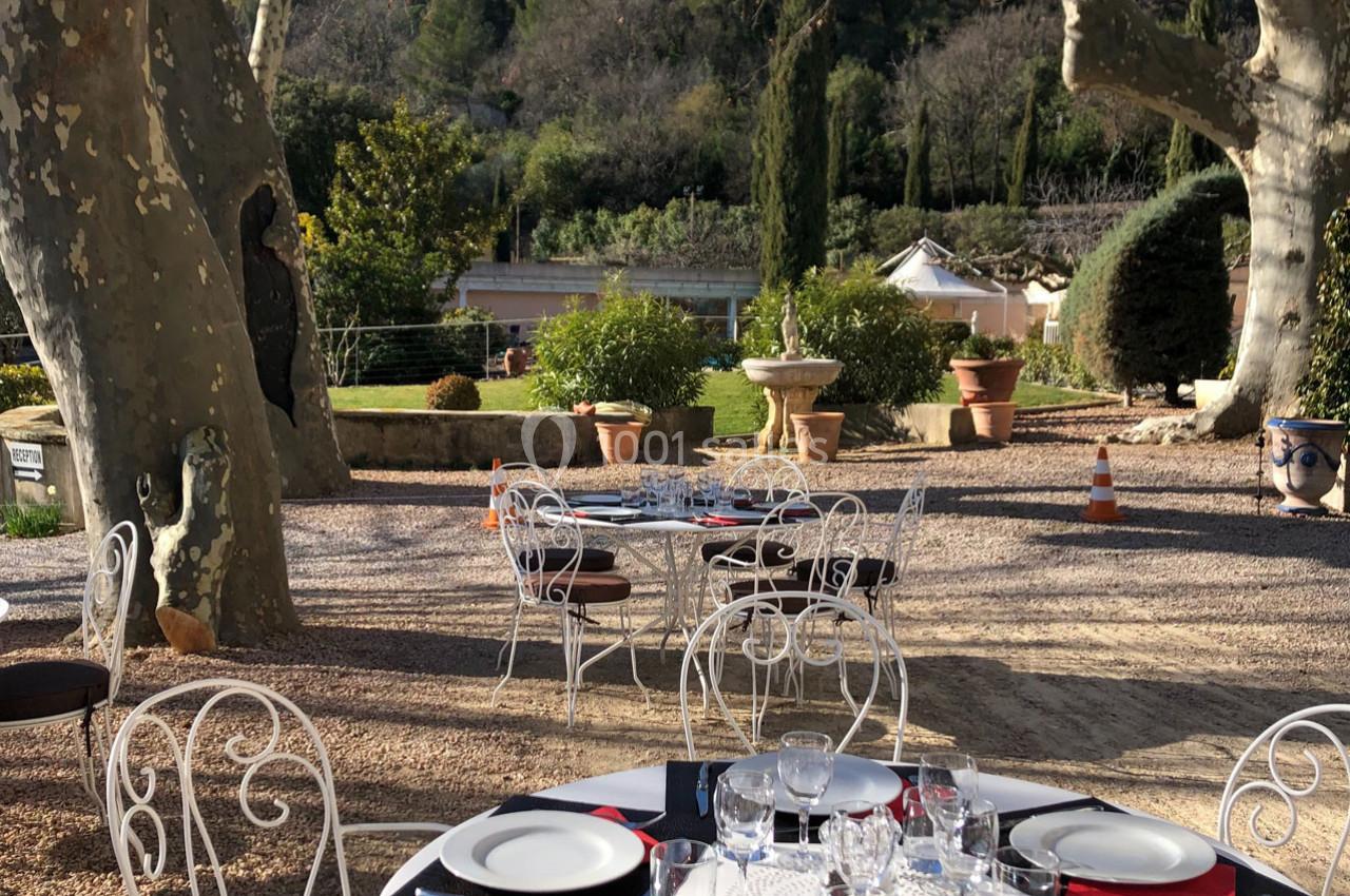 Terrasse extérieure avec tables dressées, chaises en fer forgé, arbres et vue sur un jardin en arrière-plan.