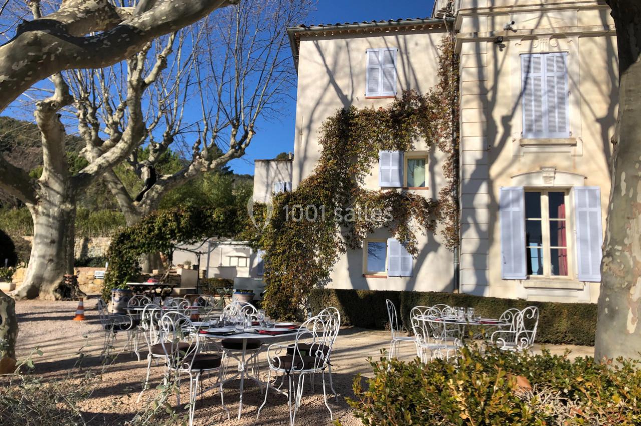 Terrasse ensoleillée avec tables et chaises en fer forgé devant une maison aux volets blancs, entourée d'arbres.
