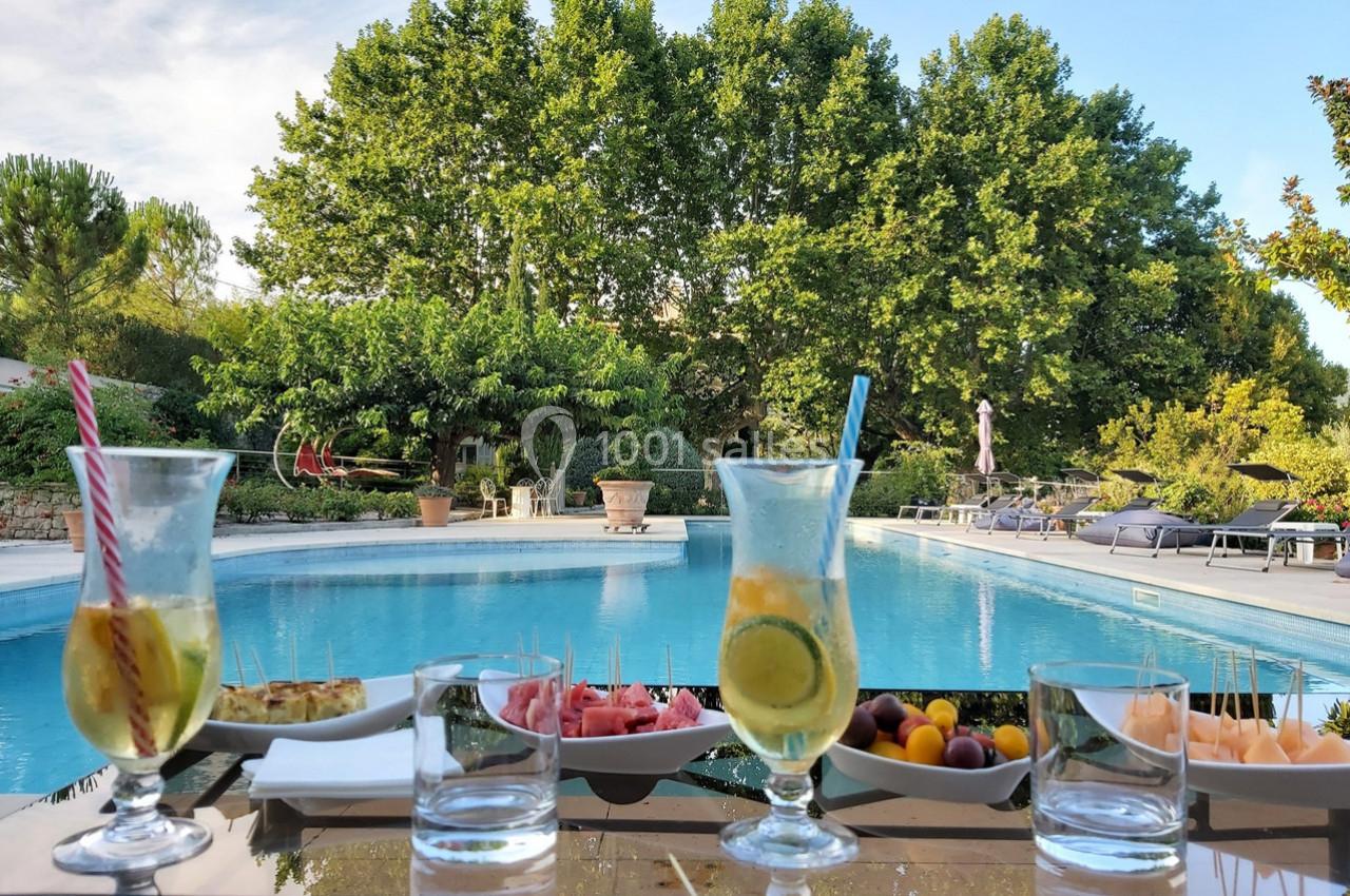 Verres de boissons et assiettes d'amuse-bouches disposés sur une table devant une piscine entourée de végétation.