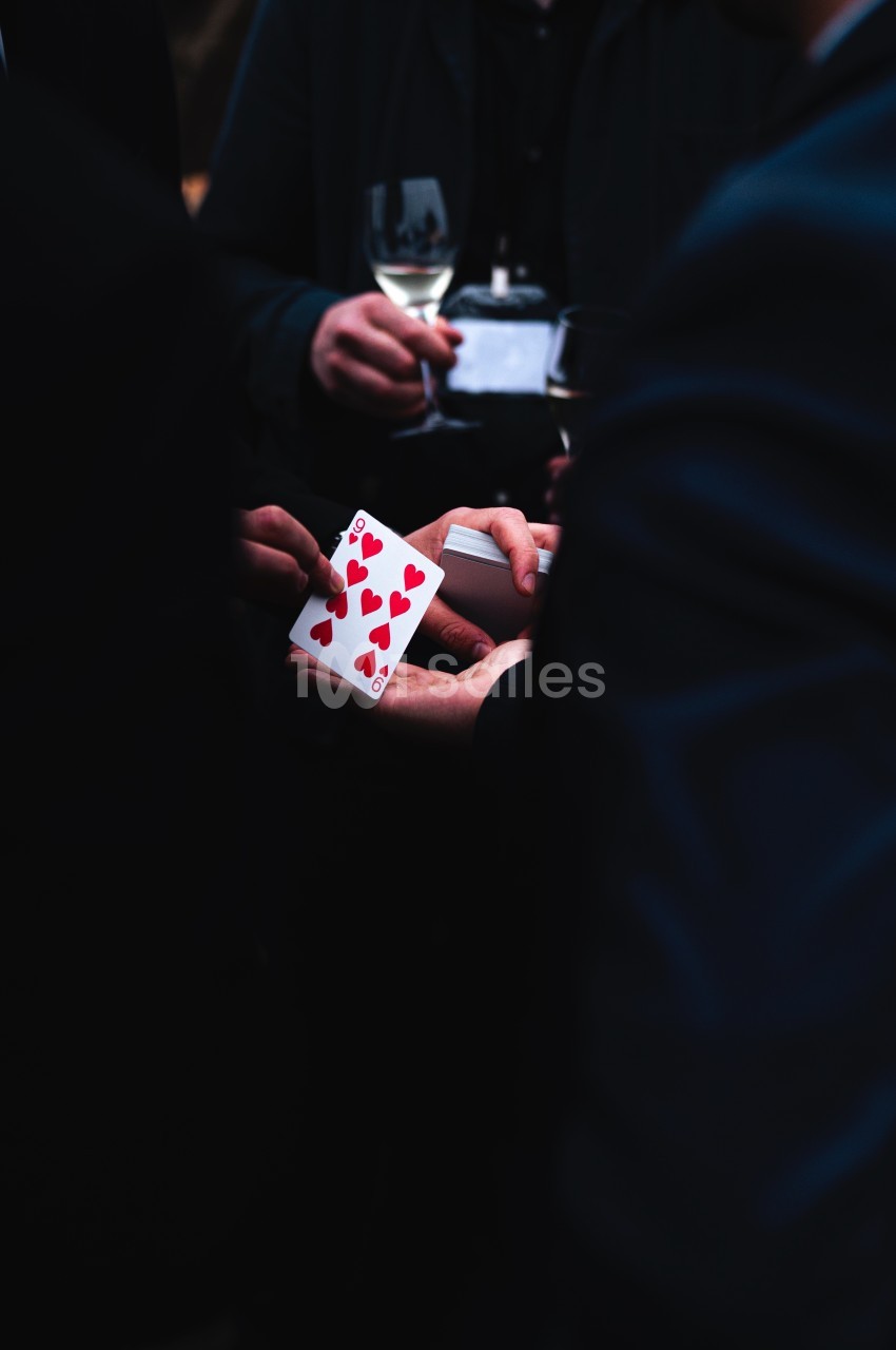 Un magicien montre une carte à jouer rouge au milieu d'un groupe de personnes tenant des verres de vin.