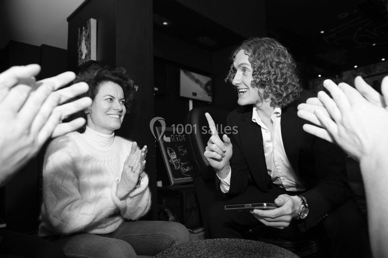 Un homme en costume parle avec animation à une femme souriante dans un café, entourés de mains applaudissant.