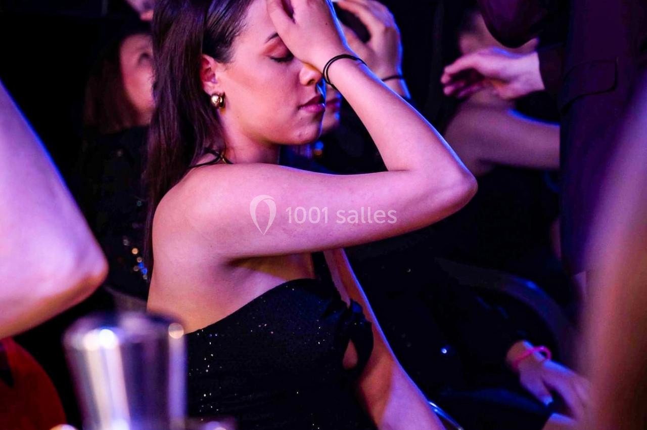 Une femme en robe noire assise, la main sur le front, dans un environnement sombre avec des lumières colorées.