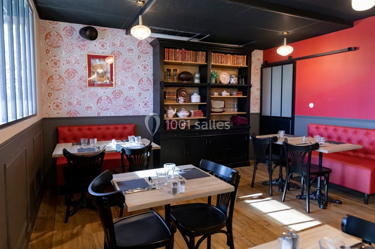 Salle de restaurant avec tables en bois, chaises noires, banquettes rouges et étagères décorées de vaisselle et livres.