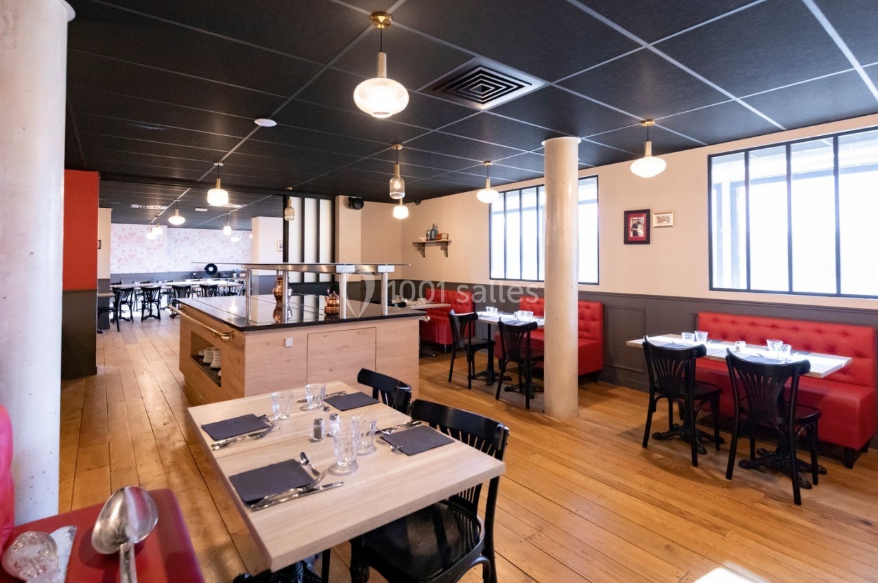Salle de restaurant lumineuse avec tables en bois, chaises noires, banquettes rouges et éclairage suspendu.