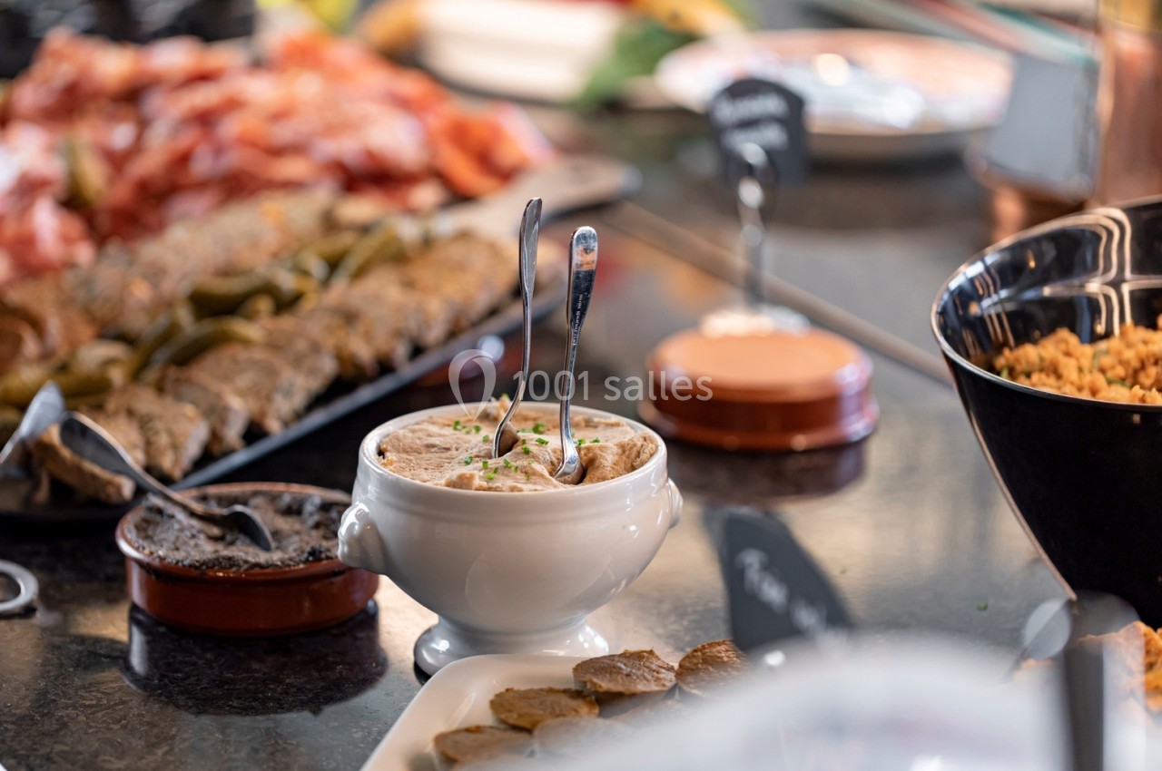 Bol de rillettes avec cuillères, entouré de charcuterie, cornichons et autres plats sur une table de buffet.