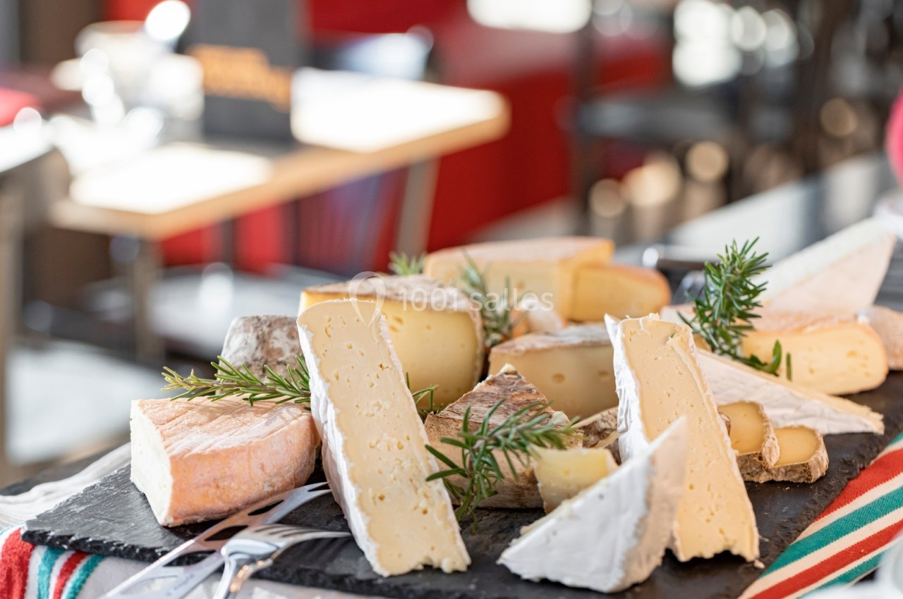 Assortiment de fromages variés disposés sur une ardoise avec des brins de romarin, en arrière-plan flou d'une salle.