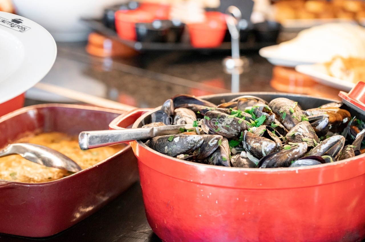 Moules cuites servies dans une marmite rouge avec une cuillère, accompagnées d'un plat en sauce à côté.