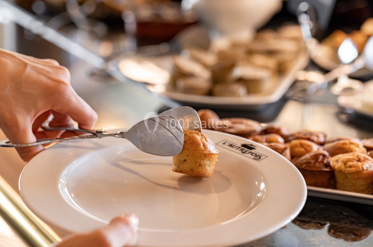 Une main utilise une pince pour déposer un petit gâteau sur une assiette blanche devant un buffet de pâtisseries.