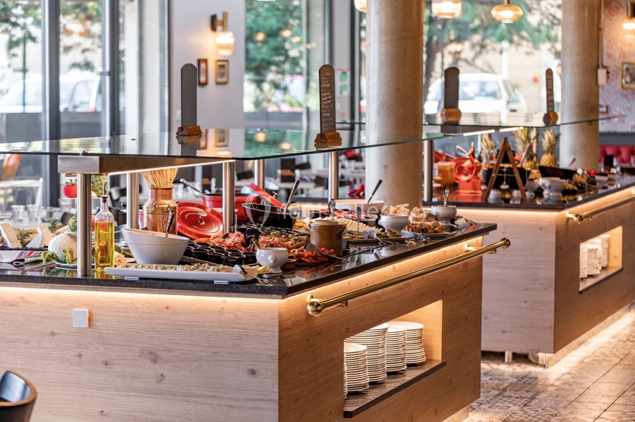 Buffet varié avec plats, condiments et ustensiles disposés sur des comptoirs en bois dans un restaurant lumineux.