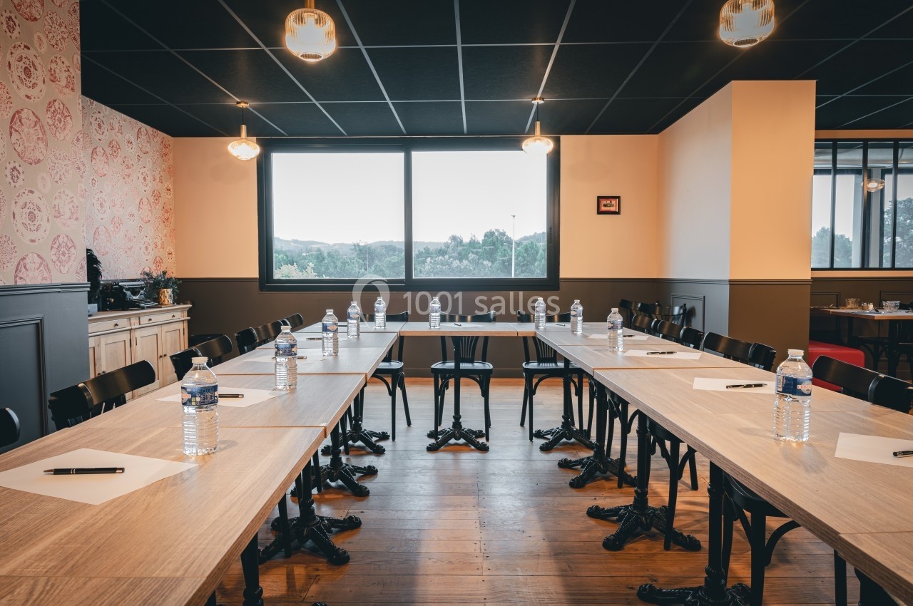 Salle de réunion lumineuse avec tables disposées en U, bouteilles d'eau, stylos et papiers sur chaque place.